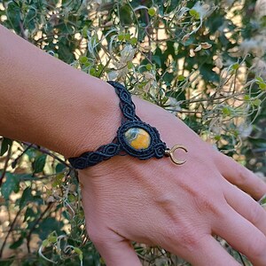 Op de afbeelding: Een zwarte macramé armband met een centrale ovaalvormige steen met gele en grijze gevlekte patronen. De armband bevat een kleine goudkleurige halve maan bedel bevestigd aan de zijkant van de steenzetting, vastgezet met geknoopt koord.