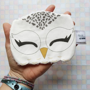 Peut inclure: Visage de hibou en tissu blanc avec des yeux noirs, un bec jaune et un motif tacheté gris et blanc sur le dessus. Le tissu est légèrement effiloché sur les bords. Le visage de hibou est tenu dans une main avec un bracelet bleu et rose.