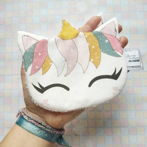 Puede incluir: Una toallita de baño blanca en forma de unicornio con una crin rosa, amarilla y verde y un cuerno dorado. La toallita tiene una cara sonriente con los ojos cerrados y corazones rosas en la parte inferior. La toallita se sostiene en una mano con una pulsera azul y verde. La toallita tiene una etiqueta que dice "Joyeux Anniversaire Ma Maman".