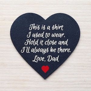 Może przedstawiać: Przedmiot w kształcie serca z ciemnoniebieskiej tkaniny z białym tekstem: "This is a shirt I used to wear. Hold it close and I'll always be there. Love, Dad." Na dole małe czerwone serce.