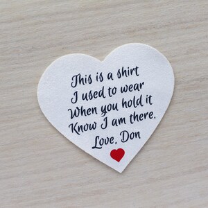 Może przedstawiać: Biała naszywka w kształcie serca z czarnym napisem: "This is a shirt I used to wear When you hold it Know I am there. Love, Don." Na dole małe czerwone serce.
