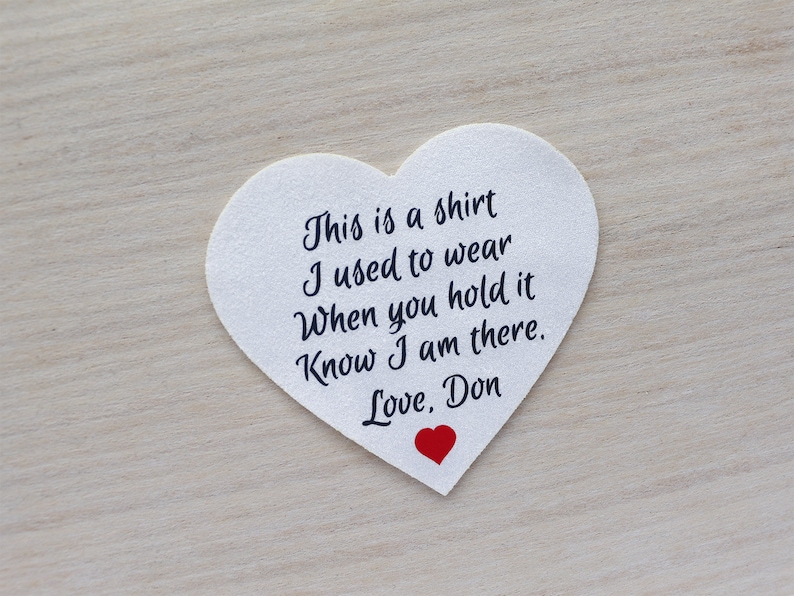 Może przedstawiać: Biała naszywka w kształcie serca z czarnym napisem: "This is a shirt I used to wear When you hold it Know I am there. Love, Don." Na dole małe czerwone serce.