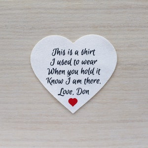 Może przedstawiać: Biała naszywka w kształcie serca z czarnym napisem: "This is a shirt I used to wear When you hold it Know I am there. Love, Don." Na dole małe czerwone serce.