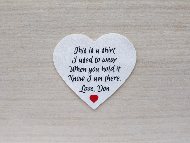 Może przedstawiać: Biała naszywka w kształcie serca z czarnym napisem: "This is a shirt I used to wear When you hold it Know I am there. Love, Don." Na dole małe czerwone serce.