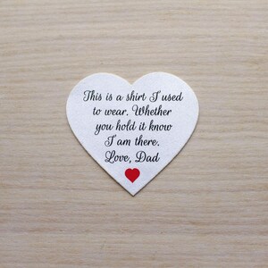 May include: A white heart-shaped item with the text "This is a shirt I used to wear. Whether you hold it know I am there. Love, Dad" in black script. A small red heart is at the bottom. The item is on a light brown wooden surface.