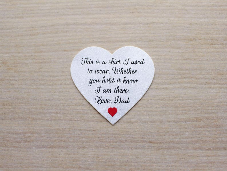 May include: A white heart-shaped item with the text "This is a shirt I used to wear. Whether you hold it know I am there. Love, Dad" in black script. A small red heart is at the bottom. The item is on a light brown wooden surface.