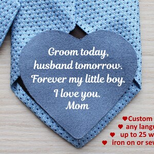 May include: A blue tie with a gray felt heart sewn on top. The heart has the text "Groom today, husband tomorrow. Forever my little boy. I love you. Mom" written in white. The text below the heart reads "Custom text, any language, up to 25 words, iron on or sew on."