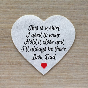 Może przedstawiać: Biały przedmiot w kształcie serca z napisem "This is a shirt I used to wear. Hold it close and I'll always be there. Love, Dad" w kolorze czarnym. Małe czerwone serce znajduje się na dole. Tło to jasne usłojenie drewna.