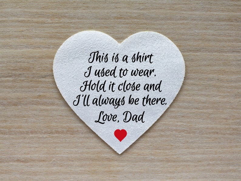 Może przedstawiać: Biały przedmiot w kształcie serca z napisem "This is a shirt I used to wear. Hold it close and I'll always be there. Love, Dad" w kolorze czarnym. Małe czerwone serce znajduje się na dole. Tło to jasne usłojenie drewna.