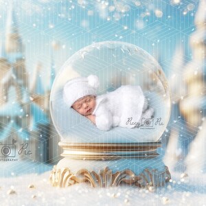 Puede incluir: Un bebé recién nacido durmiendo, vestido con un conjunto y gorro de punto blanco, dentro de una bola de nieve. La bola de nieve se asienta sobre una base dorada decorativa, con un fondo borroso de un país de las maravillas invernal.