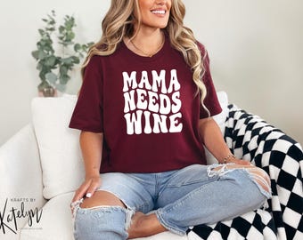 Puede incluir: Una camiseta de color burdeos con el texto "Mama Needs Wine" en blanco, con letras de estilo retro.