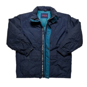 May include: A navy blue Fila jacket with a teal lining and a zipper closure. The jacket has a hood and two pockets on the front. The Fila logo is visible on the left chest and the zipper pull.