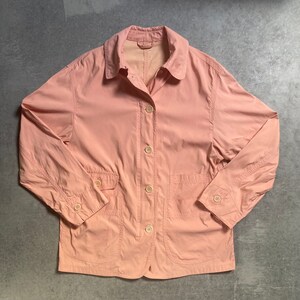 Puede incluir: Una chaqueta de trabajo rosa claro con cierre de botones y varios bolsillos.