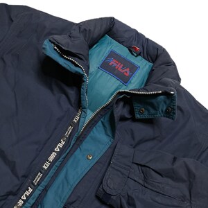 May include: A navy blue jacket with a teal lining and a Fila Gore-Tex label. The jacket has a zipper closure and a snap button closure. The Fila logo is visible on the inside of the collar.