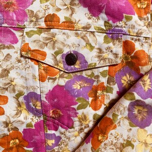 May include: A close-up of a white denim jacket with a floral print. The print features pink, purple, and orange flowers on a white background. The jacket has a pocket on the left side with a button closure.