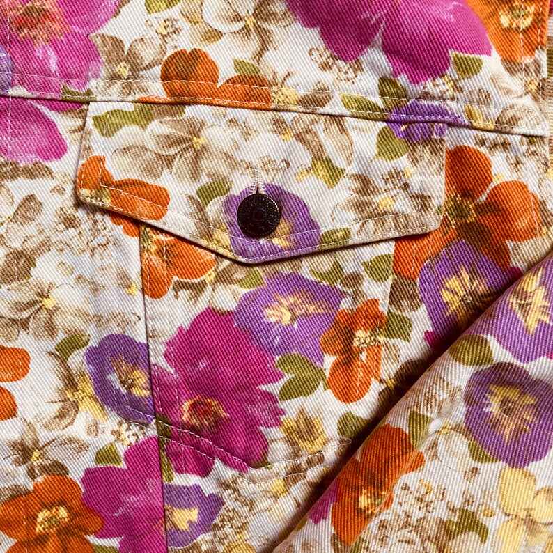 May include: A close-up of a white denim jacket with a floral print. The print features pink, purple, and orange flowers on a white background. The jacket has a pocket on the left side with a button closure.