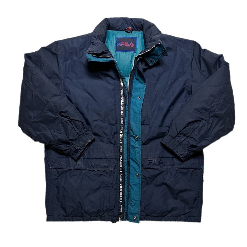 May include: A navy blue Fila jacket with a teal lining and a zipper closure. The jacket has a hood and two pockets on the front. The Fila logo is visible on the left chest and the zipper pull.