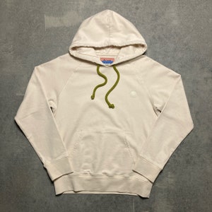 May include: A cream-colored hooded sweatshirt with a green drawstring and a small white logo on the front. The logo is a circle with a small circle inside it. The sweatshirt has a kangaroo pocket on the front.