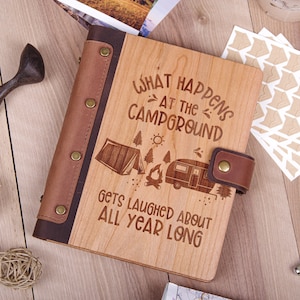May include: A wooden journal with a brown leather cover and a quote that reads "What happens at the campground gets laughed about all year long". The journal has a camping scene engraved on the front cover, including a tent, a campfire, and a camper.