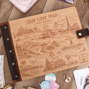May include: A rectangular wooden photo album with a dark brown leather spine secured by four metal rivets and a matching leather strap closure. The cover features an engraved map titled Our Love Map, Kate and Alex, with illustrations of landmarks, a dog, and text.