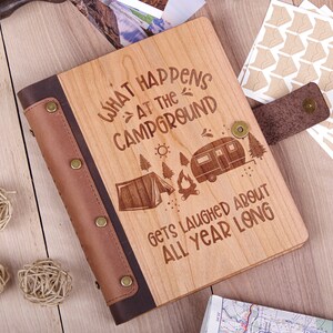 May include: A wooden photo album with a brown leather cover and a design that says "What happens at the campground gets laughed about all year long". The design includes a tent, a campfire, and a camper.