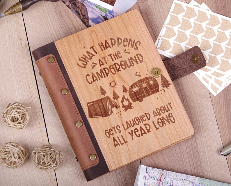 May include: A wooden photo album with a brown leather cover and a design that says "What happens at the campground gets laughed about all year long". The design includes a tent, a campfire, and a camper.