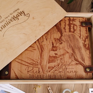 May include: A wooden box with a lid that opens to reveal a personalized engraved wooden photo album. The album features a couple's names, "Rick & Brittany", and the text "Our Greatest Adventures". The lid of the box is engraved with the text "Happy First Wedding Anniversary".