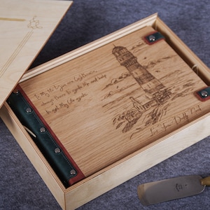May include: A wooden box with a hinged lid, containing a leather-bound journal with a lighthouse design and the inscription "To my the you are Cothea, always there to guide me and help to set my Life a-go." The box is made of light wood and has a green leather strap with metal rivets. The journal is closed and has a green leather spine with metal rivets. The box is open and the journal is visible inside. The box is sitting on a gray surface.