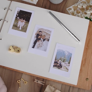 May include: A white wedding album with a wooden cover. The album is open to a page with three photos of a wedding couple and their wedding party. The photos are in a Polaroid style frame. There are two gold heart-shaped lockets and two gold keys on the page.