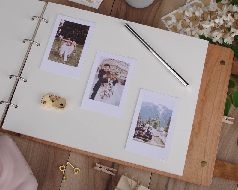 May include: A white wedding album with a wooden cover. The album is open to a page with three photos of a wedding couple and their wedding party. The photos are in a Polaroid style frame. There are two gold heart-shaped lockets and two gold keys on the page.