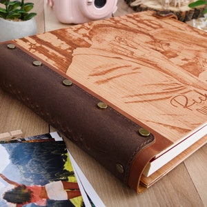 May include: A wooden photo album with a brown leather cover and brass accents. The album has a personalized inscription that reads "Rick is our greatest adventure." The album is closed and lying on a wooden surface.
