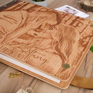 May include: A wooden photo album with a brown leather strap and metal accents. The cover features a laser-engraved image of a couple with the text "Dick & Brittany, Greatest Adventures".
