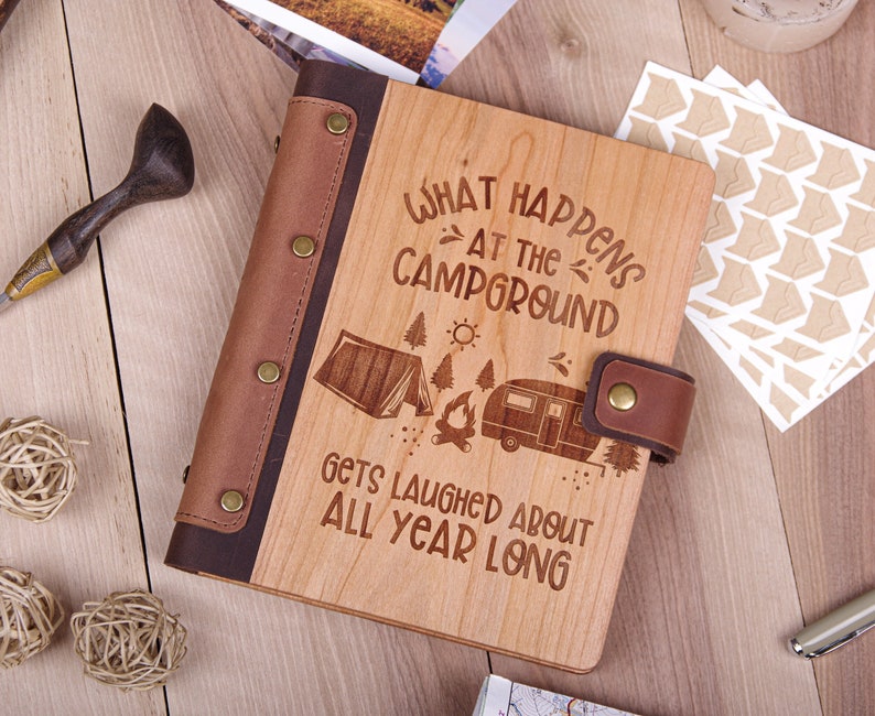 May include: A wooden journal with a brown leather cover and a quote that reads "What happens at the campground gets laughed about all year long". The journal has a camping scene engraved on the front cover, including a tent, a campfire, and a camper.