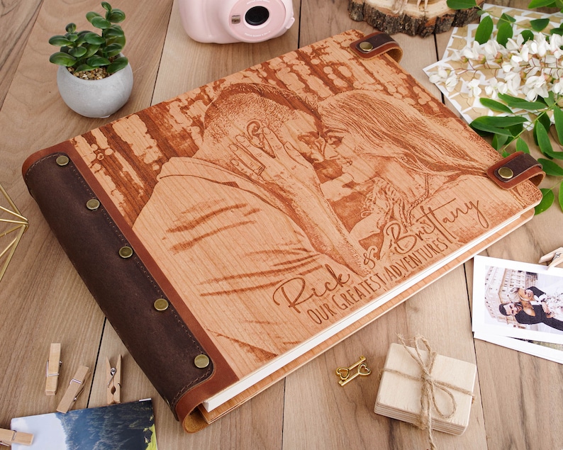 May include: A wooden photo album with a brown leather cover and a personalized inscription that reads "Rick & Brittany Our Greatest Adventures." The album features a laser-engraved image of a couple embracing.