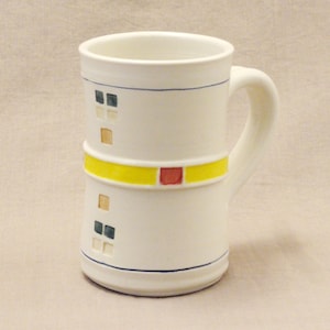 May include: White ceramic mug with a yellow, red, and blue geometric design. The mug has a handle and is decorated with a band of yellow and red squares.