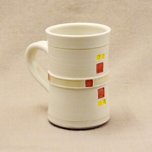 May include: White ceramic mug with a red and yellow geometric design. The mug has a rounded handle.