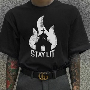 Op de afbeelding: Zwart T-shirt met een witte afbeelding van een brandende kerk met de tekst "Stay Lit". De persoon die het shirt draagt heeft een zwarte riem met een gouden gesp en een grote tatoeage op zijn arm.