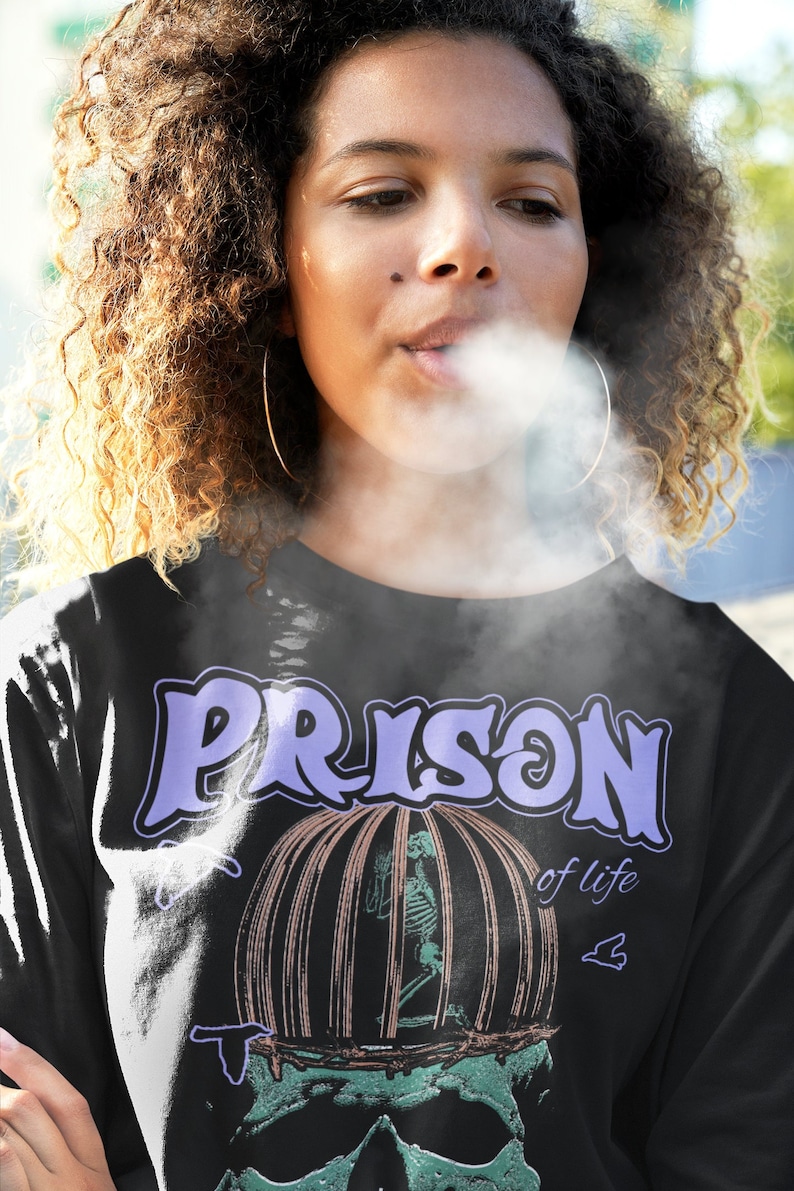 May include: A black long-sleeve shirt with a graphic print of a skull with a cage on top. The text "PRISON of life" is printed on the shirt.
