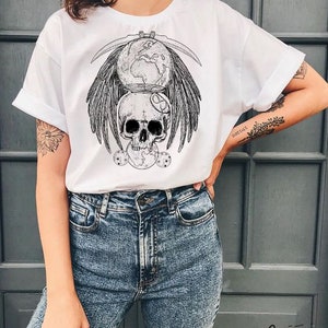 May include: White t-shirt with a black and white graphic print of a skull with wings, a globe, and a crescent moon. The graphic is centered on the shirt.