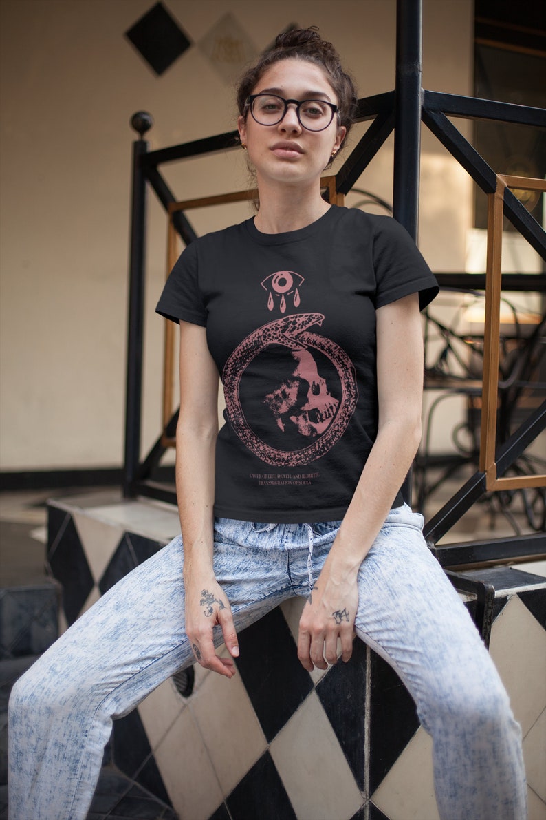 以下が含まれることがあります： ピンクのグラフィックデザインが施された黒いTシャツ。グラフィックには、蛇が巻き付いた頭蓋骨が描かれており、ピンクの目と3つのピンクの涙も含まれています。グラフィックの下には、「UNTIL THE DAY, DEATH AND ALL ELSE TRANSGRESS YOUR SOUL, IT IS A」というテキストが印刷されています。