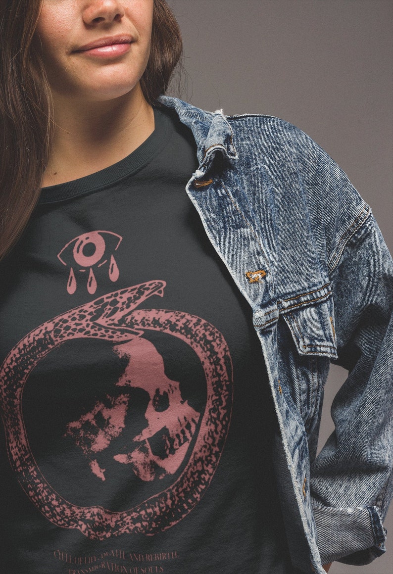 以下が含まれることがあります： 黒いTシャツに、ピンク色のグラフィックデザインが施されています。デザインは、蛇の中に頭蓋骨が描かれており、その上に泣いている目が描かれています。グラフィックの下には、「CYCLE OF DEATH AND REBIRTH TRANSFORMATION OF SOULS」というテキストが書かれています。