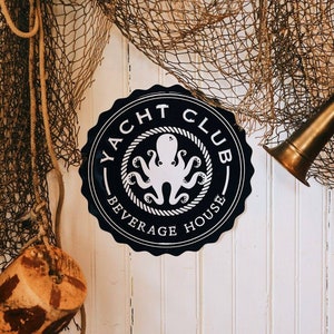 May include: A black and white metal sign with a nautical theme. The sign reads "Yacht Club Beverage House" and features an octopus with eight tentacles.