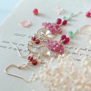 Könnte beinhalten: Elegante goldfarbene Ohrringe mit Anhängern aus rosa und roten Edelsteinen, ergänzt durch klare und grüne Akzente. Die Ohrringe haben ein zartes, fließendes Design, perfekt, um einen Hauch von Raffinesse zu verleihen. Auf einem Buch platziert.