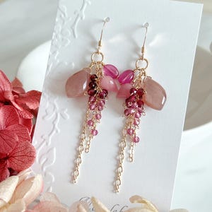Peut inclure: Boucles d'oreilles pendantes avec des pierres précieuses roses et bordeaux. Les boucles d'oreilles présentent un fil et une chaîne dorés, avec un mélange de pierres facettées et lisses. Les boucles d'oreilles sont présentées sur une carte blanche.
