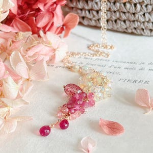 Könnte beinhalten: Eine zarte goldene Halskette mit einem Cluster aus rosa und roten Edelsteinen. Der Anhänger hat eine Mischung aus Rubin und anderen rosa Steinen mit einer zarten Kette. Die Halskette wird mit rosa Blumen und einem Vintage-Buch präsentiert.