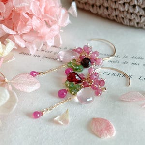 May include: Gold-coloured hoop earrings adorned with clusters of pink, green, and dark red gemstones. The earrings have dangling chains with pink beads. Soft pink flower petals are scattered around on a light-coloured surface.