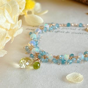 Könnte beinhalten: Ein zartes goldfarbenes Armband mit Clustern aus blauen, weißen und gelben Edelsteinen. Das Armband hat einen Anhänger mit einem großen gelben tropfenförmigen Stein und einem kleinen grünen runden Stein. Die Edelsteine sind mit Golddraht aufgereiht.