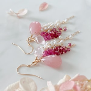 Peut inclure: Une paire de boucles d'oreilles pendantes avec des crochets dorés, de grosses pierres facettées en forme de goutte rose pâle, des grappes de petites perles d'eau douce blanches et des perles de pierres précieuses rose foncé en cascade.