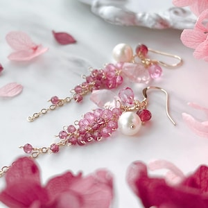 Può includere: Orecchini pendenti con pietre preziose rosa e perle, sospesi a ganci dorati. Gli orecchini presentano una cascata di tonalità rosa, dal chiaro allo scuro, con alcune pietre chiare e sfaccettate. Design delicato e femminile.