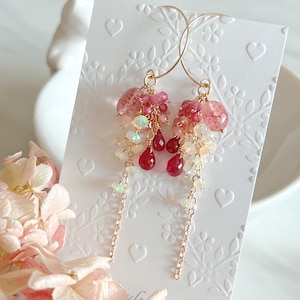May include: Gold-tone hoop earrings with clusters of pink and red gemstones. The earrings feature a mix of round and teardrop-shaped stones, with delicate chain accents. The background is a white card with embossed floral and heart patterns.